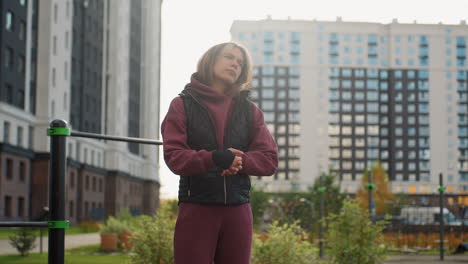 calm focused woman standing beside outdoor exercise bars under bright sunlight rolling hands then gently rotating neck in smooth motion to warm muscles for fitness routine in urban park setting