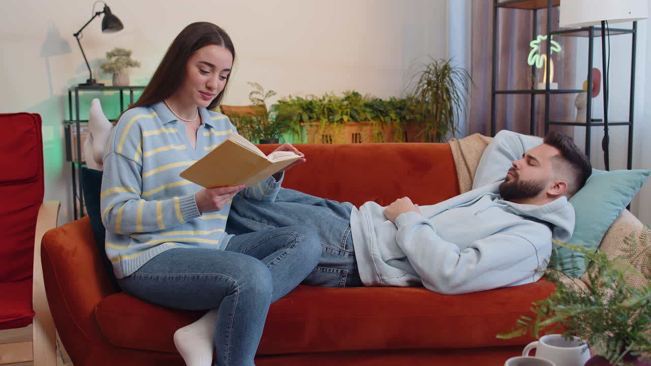 Woman reading story from book while displeased tired boyfriend man falling asleep snoring at home