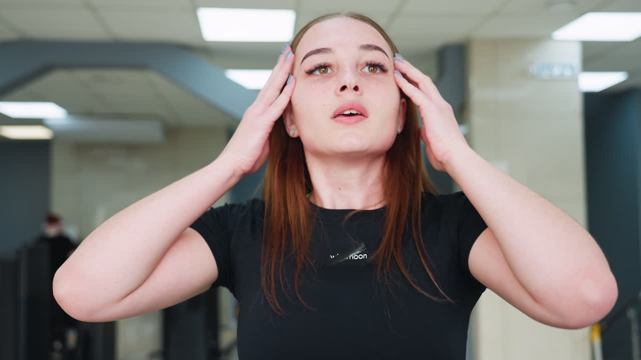resilient woman in black activewear adjusts hair back while breathing heavily after workout inside modern gym with slightly blurred background featuring workout equipment and tiled ceiling