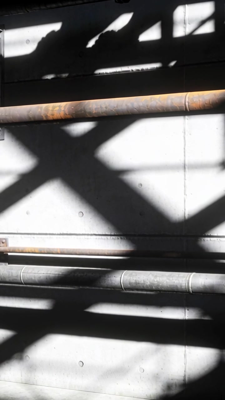 Vertical video: Shifting camera revealing lattice shadows moving across pale wall with two pipes