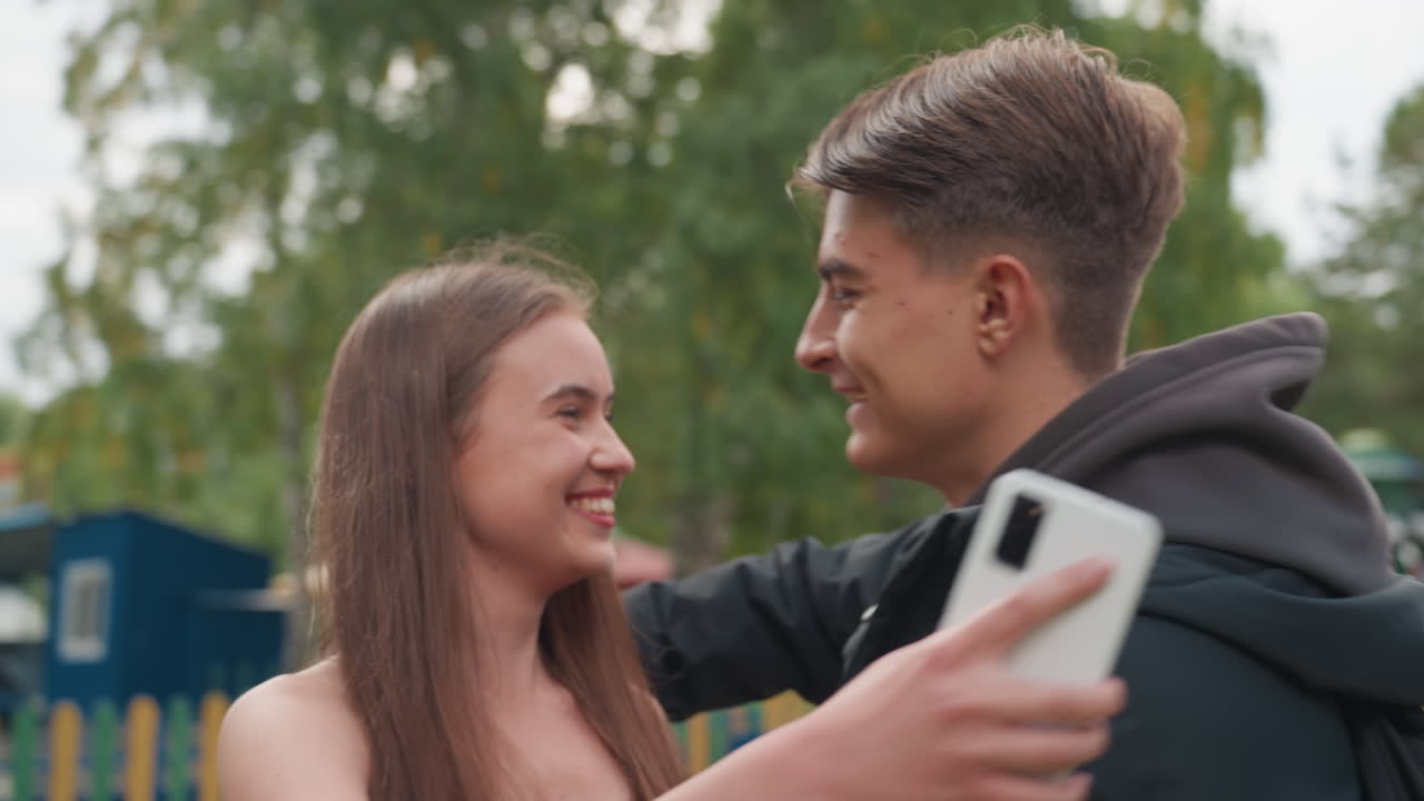 Un giovane sorprende una giovane donna con un abbraccio sentito in un parco divertimenti all'aperto vicino a una giostra e ad alcuni alberi, felpa con cappuccio e vestito casual, sorrisi emozionanti, caloroso momento di ricongiungimento, inquadratura cinematografica spontanea, luce diurna
