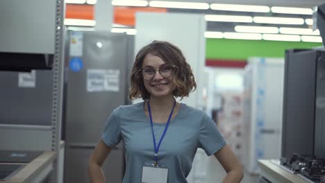 retrato positivo de una vendedora o asistente de una tienda en una tienda de supermercado. mujer con camisa azul y insignia vacía mirando a la cámara y sonriendo. electrodomésticos en el fondo