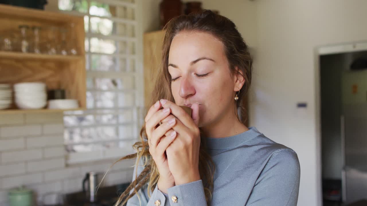 Mulher caucasiana feliz em pé na cozinha de chalé apreciando beber chá e sorrindo.