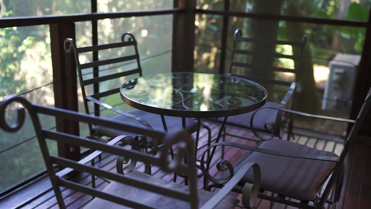 Cozy hotel room balcony featuring elegant iron furniture, including a round glass table and four chairs, overlooks lush greenery, providing a serene and inviting outdoor space