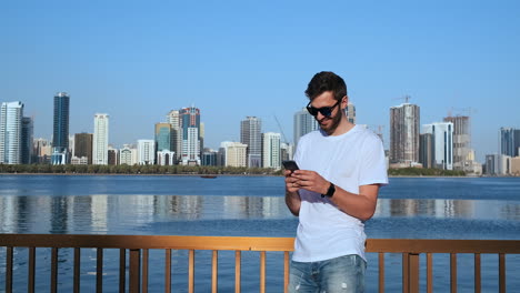 Hombre-Enviando-Mensajes-De-Texto-Usando-Una-Aplicación-En-Un-Teléfono-Inteligente-En-La-Ciudad.-Apuesto-Joven-Hombre-De-Negocios-Usando-Un-Teléfono-Inteligente-Sonriendo-Feliz-Usando-Chaqueta-De-Traje-Al-Aire-Libre.-Profesional-Masculino-Urbano-De-Unos-20-Años