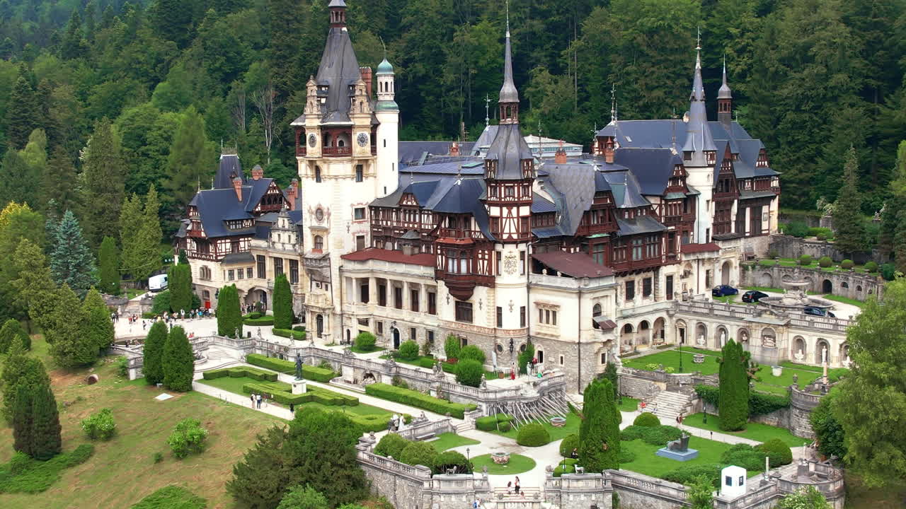 Luftfoto med drone af Peleș-slottet i Rumænien. Slot med haver og turister i Karpaterne, frodig skov omkring.