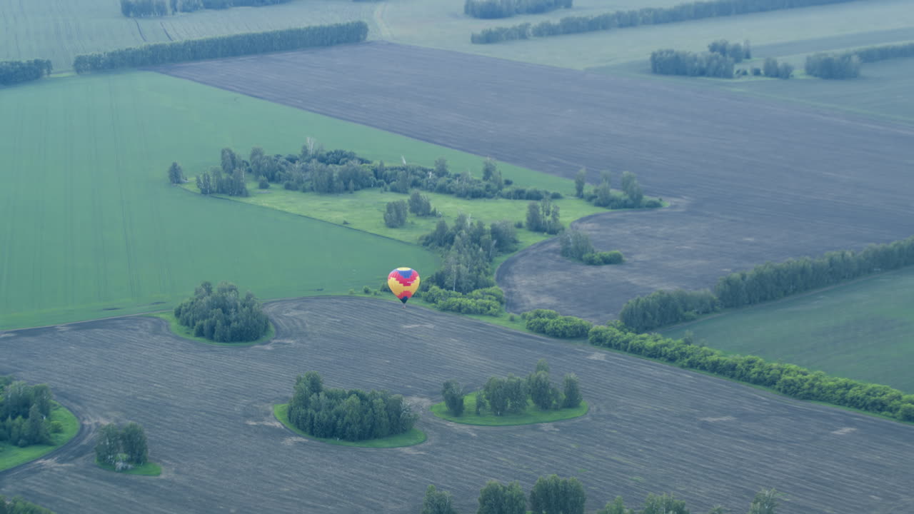 Distant view of colourful hot air balloon drifting over vast tilled and grassy fields dotted with small tree clusters under soft evening light conveying serene aerial skyward journey