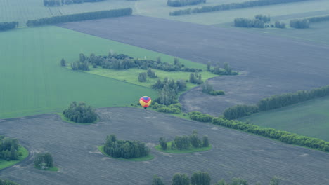 distant view of colourful hot air balloon drifting over vast tilled and grassy fields dotted with small tree clusters under soft evening light conveying serene aerial skyward journey