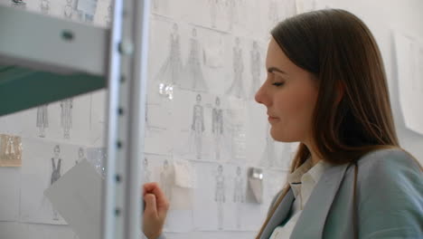 female fashion designer looking at drawings and sketches that are pinned to the wall behind her desk. studio is sunny. personal computer colorful fabrics sewing items are visible.