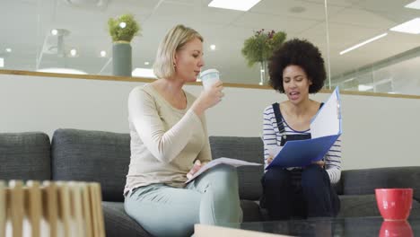 diverse female colleagues sitting on sofa, drinking coffee, having business talk in office