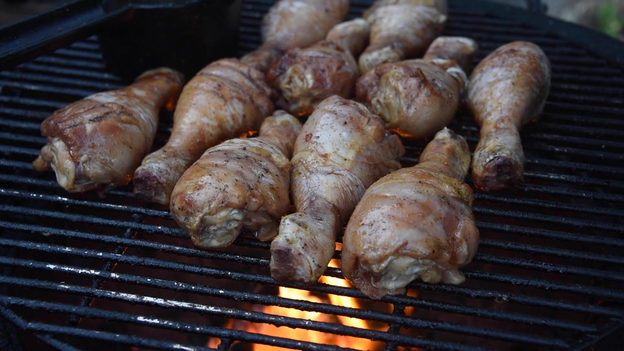 Primo piano di cosce di pollo condite che affumicano e cuociono su una griglia calda. Le fiamme lambiscono la carne mentre arrostisce lentamente all'aperto