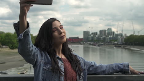 beautiful latina having fun while taking a selfie with her phone, posing and having a great time while standing on a bridge with a view of london