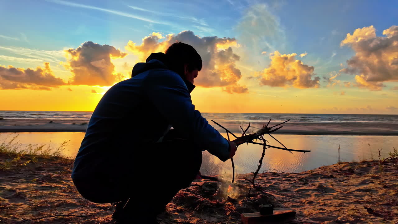 Man is making a fire on the beach while sunset, slowmotion, beautiful colors and clouds on the sky with copy space
