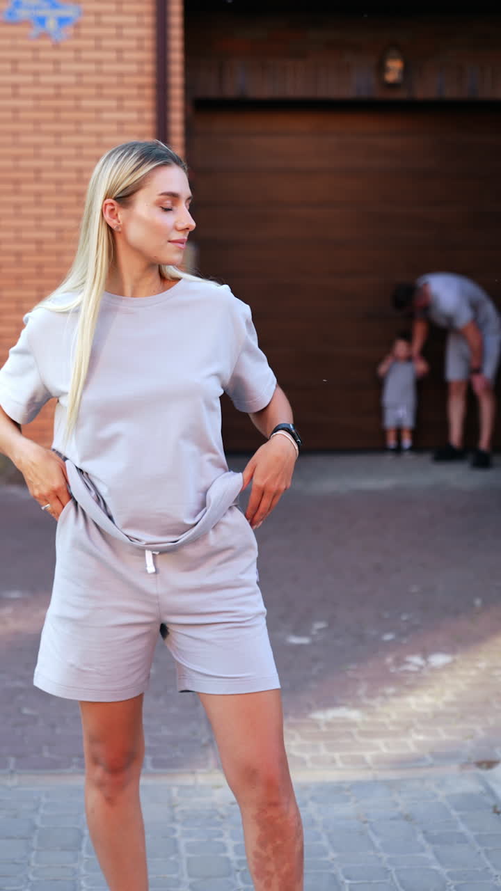 Beautiful blonde Caucasian woman wearing grey t-shirt and shorts stands outdoors. Lady pulls up her shorts and puts hands to the pockets. Man and kid at backdrop. Vertical video