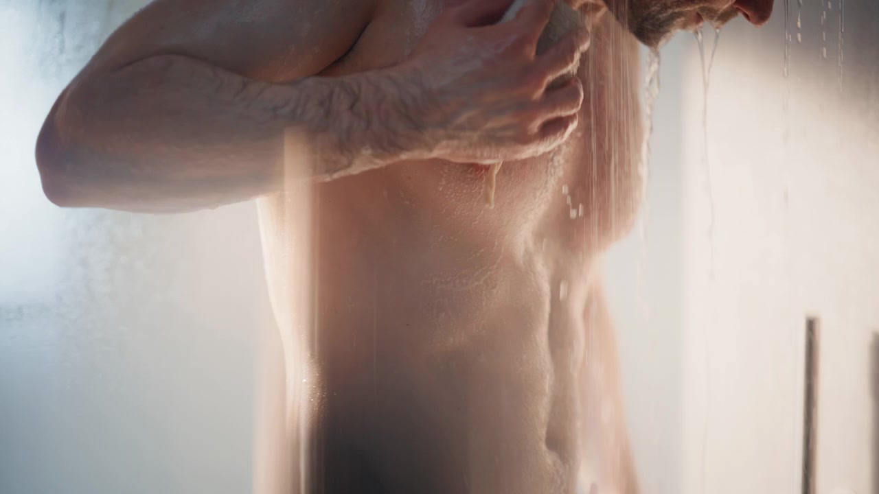 Primo piano di un uomo che si lava il corpo con una spugna di luffa sotto la doccia. Torso bagnato in un bagno fumante.
