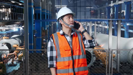 ingeniero asiático con casco de seguridad hablando en teléfono inteligente mientras está de pie en una fábrica de fabricación de turbinas eólicas
