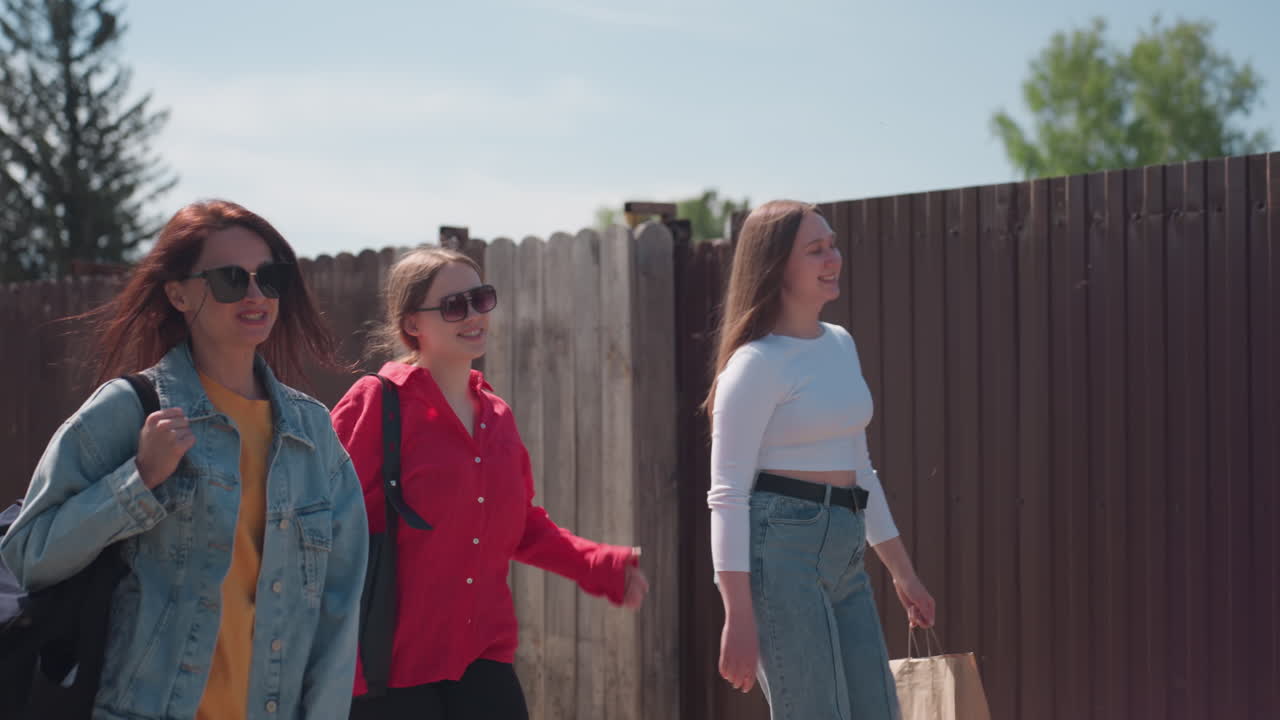 Three young women walking joyfully along rough village road on sunny day, chatting and laughing together, one holding shopping bag, surrounded by greenery, wooden fences, and utility poles