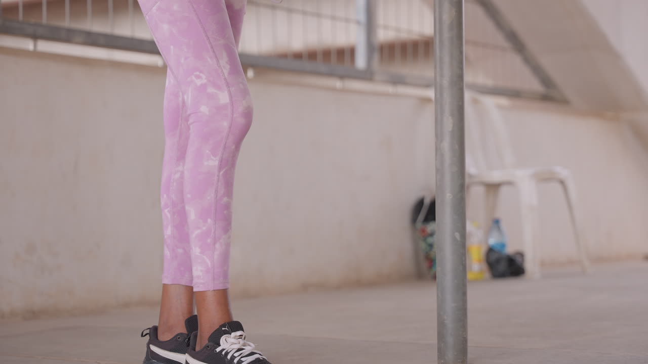 Primo piano di gambe in leggings rosa, persona che si stira accanto a un palo metallico su un balcone di cemento, scarpe da ginnastica nere, sedia di plastica e borsa sullo sfondo, luce naturale soffusa, atmosfera di allenamento urbano casual.