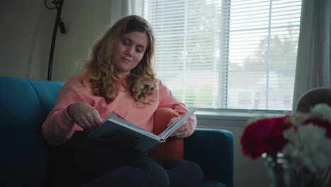 woman in pink sweater sits on blue couch reading through pages of a book by a window in the daytime