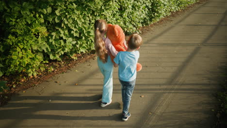 Back view of boy and girl walking together on paved path bordered by bushes, with girl holding large teddy bear and boy adjusting its head, soft shadows of background elements cast across ground