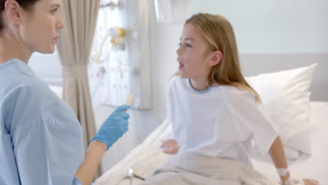 caucasian female doctor in gloves with spatula checking throat of girl hospital patient, slow motion