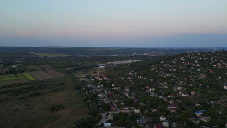 Pueblo-En-La-Cima-De-Una-Colina-Inmerso-En-Una-Exuberante-Vegetación:-El-Vuelo-Nocturno-De-Un-Dron-Sobre-Un-Pueblo-Verde,-Bañado-Por-Una-Puesta-De-Sol-Azul-Rosado,-Un-Vuelo-Panorámico-Tranquilo