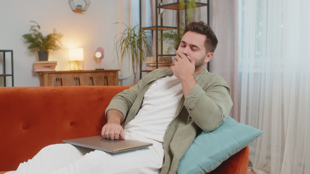 Stressed young male freelancer sitting on sofa throwing papers while using laptop computer at home