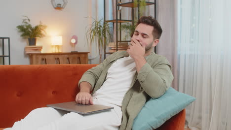 stressed young male freelancer sitting on sofa throwing papers while using laptop computer at home