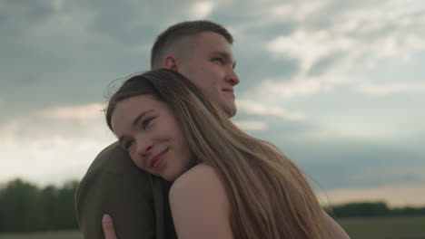 closeup of woman embracing man warmly in open field under dramatic cloudy sky, long hair flowing over shoulder, eyes closed in gentle smile, soft golden light illuminating affectionate bond