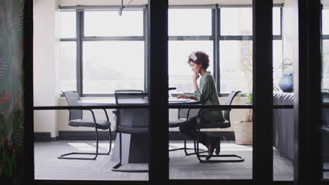 young black businesswoman working alone in an office, seen through glass wall, full length, zoom out