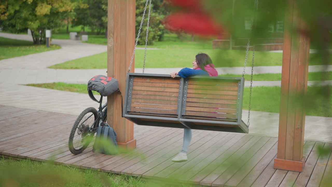 woman sitting on swing bench beside bicycle, pink jacket, helmet on handlebar, backpack resting against wooden post, gazing across quiet park with green trees and boardwalk, contemplative mood, gentle