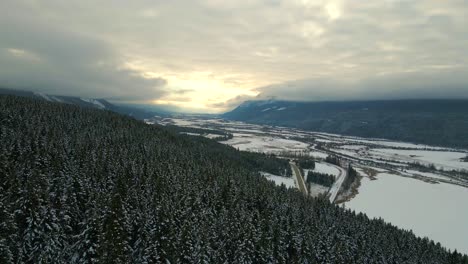 Golden-Hour-Aerial-View-of-Snowy-Forest-and-Cloud-Covered-Mountain-Scenery-in-British-Columbia,-Canada:-Stunning-Winter-Landscape-from-Drone's-Perspective