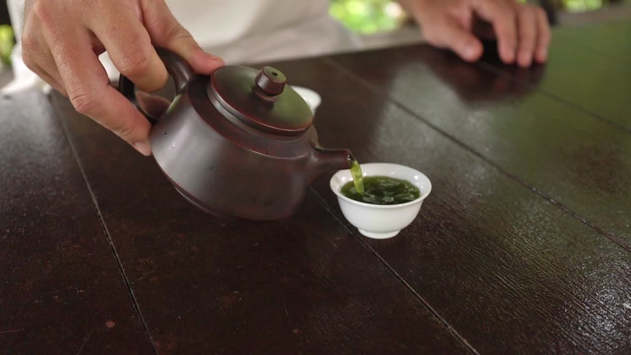 Un primo piano di una persona che versa matcha verde da una tradizionale teiera di argilla scura in piccole tazzine da tè di porcellana bianca su un tavolo di legno