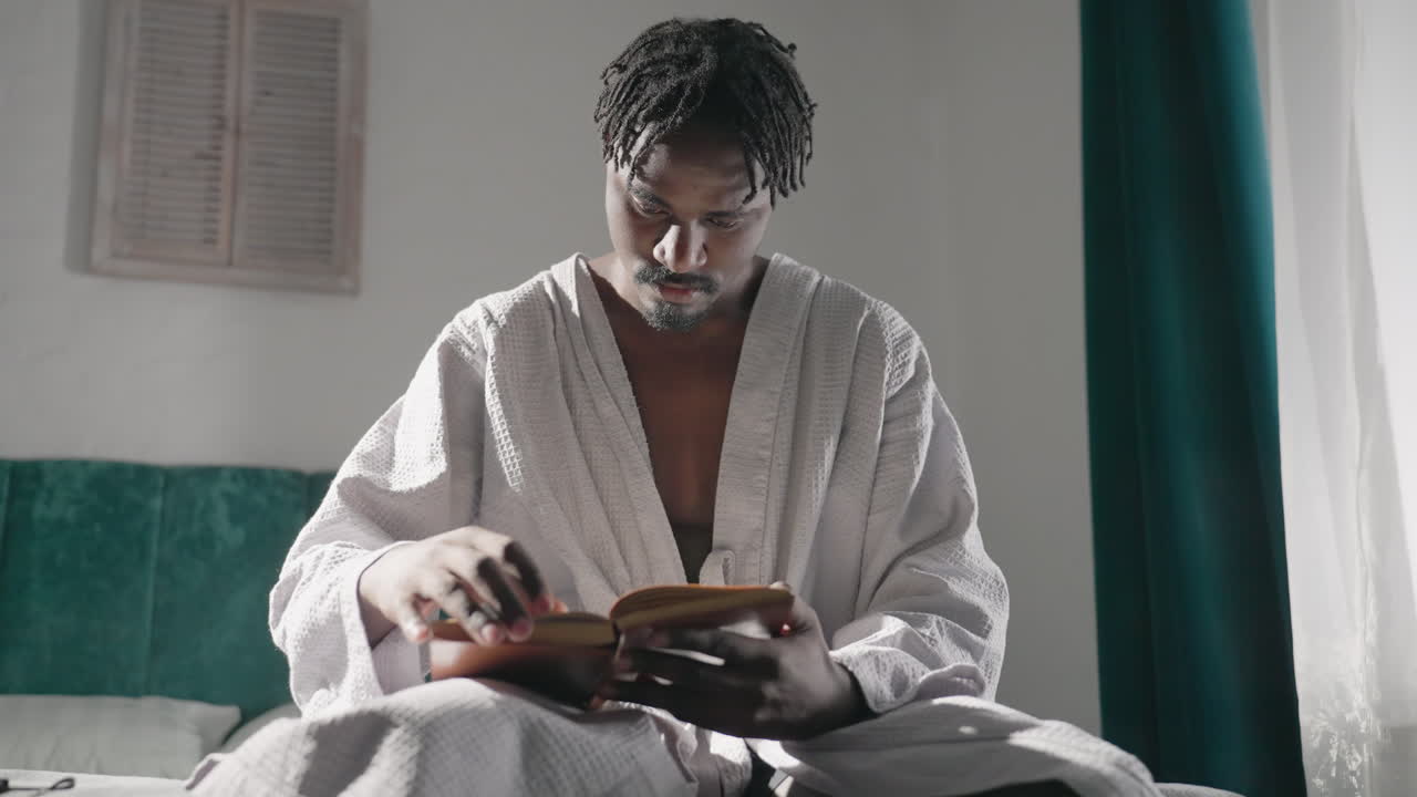Guy in bathrobe sitting on bed, reading a book in soft morning light. Quiet, focused expression. Relaxing, peaceful atmosphere in cozy room with sunlight streaming through window