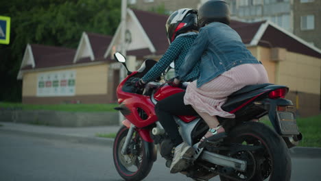 two women ride a power bike, with the passenger holding onto the rider as they travel down an urban road, buildings and pedestrians are visible in the background