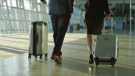 Back-View-On-The-Businessman-And-Businesswoman-Walking-The-Airport-Hall-And-Carrying-Their-Suitcases-On-The-Wheels-Before-The-Departure-To-The-Working-Trip