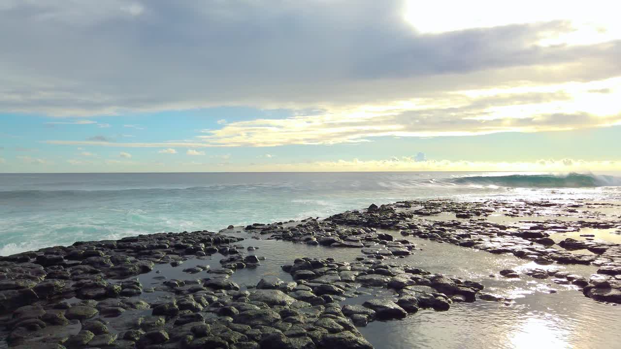4K Hawaii Kauai boom up and pan left from waves crashing in distance, grass at edge of pools on lava rock in foreground to ocean waves building and crashing on lava rock with bright partly cloudy sky