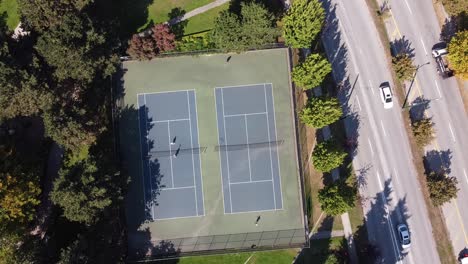 2-2-Luftaufnahme-Aus-Der-Vogelperspektive-Von-Paaren,-Die-Tennis-Spielen,-In-Einem-Modernen-Park-Neben-Einem-Luxusclub-In-Der-Nähe-Einer-Gartenstraßenautobahn-Mit-Möwen-Und-Tauben,-Die-über-Die-Athletischen-Erwachsenen-Fliegen,-Die-Sich-Am-Netz-Treffen