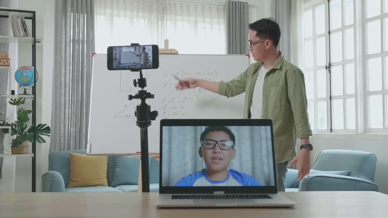 Asian Male Teacher With Glasses Shoots Video By Smartphone Camera And Having Video Call On Laptop While Teaching Math At Home