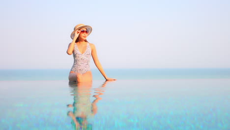 a woman sitting on the edge of a swimming pool reaches up to adjust her sunglasses