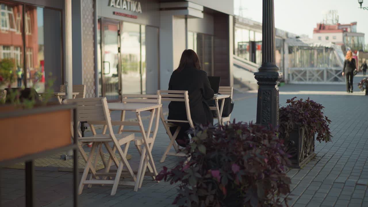 Back view of professional lady typing on laptop outdoors at cafeteria, partial view of someone in black outfit walking in the distance, with plants and wooden chairs surrounding the area