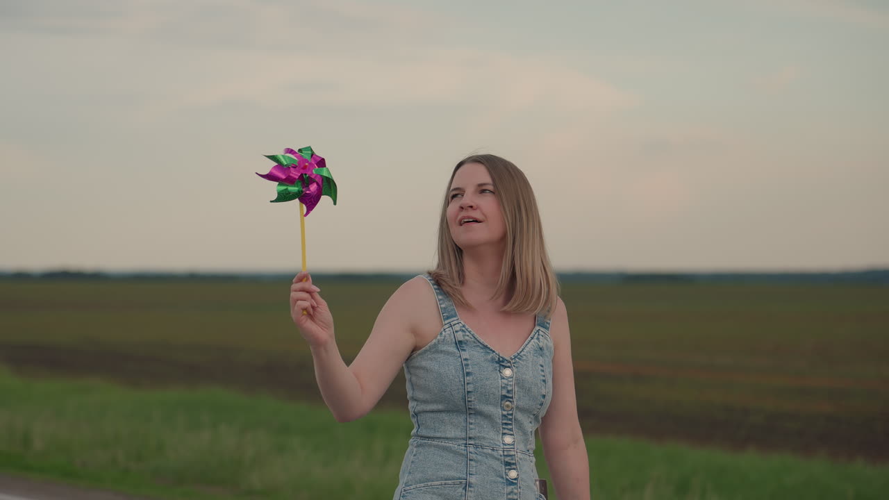 Woman walking beside grassy field blows pinwheel in hand to set it spinning under overcast sky wind swirling hair capturing playful energy carefree motion in bright summer outdoors scene