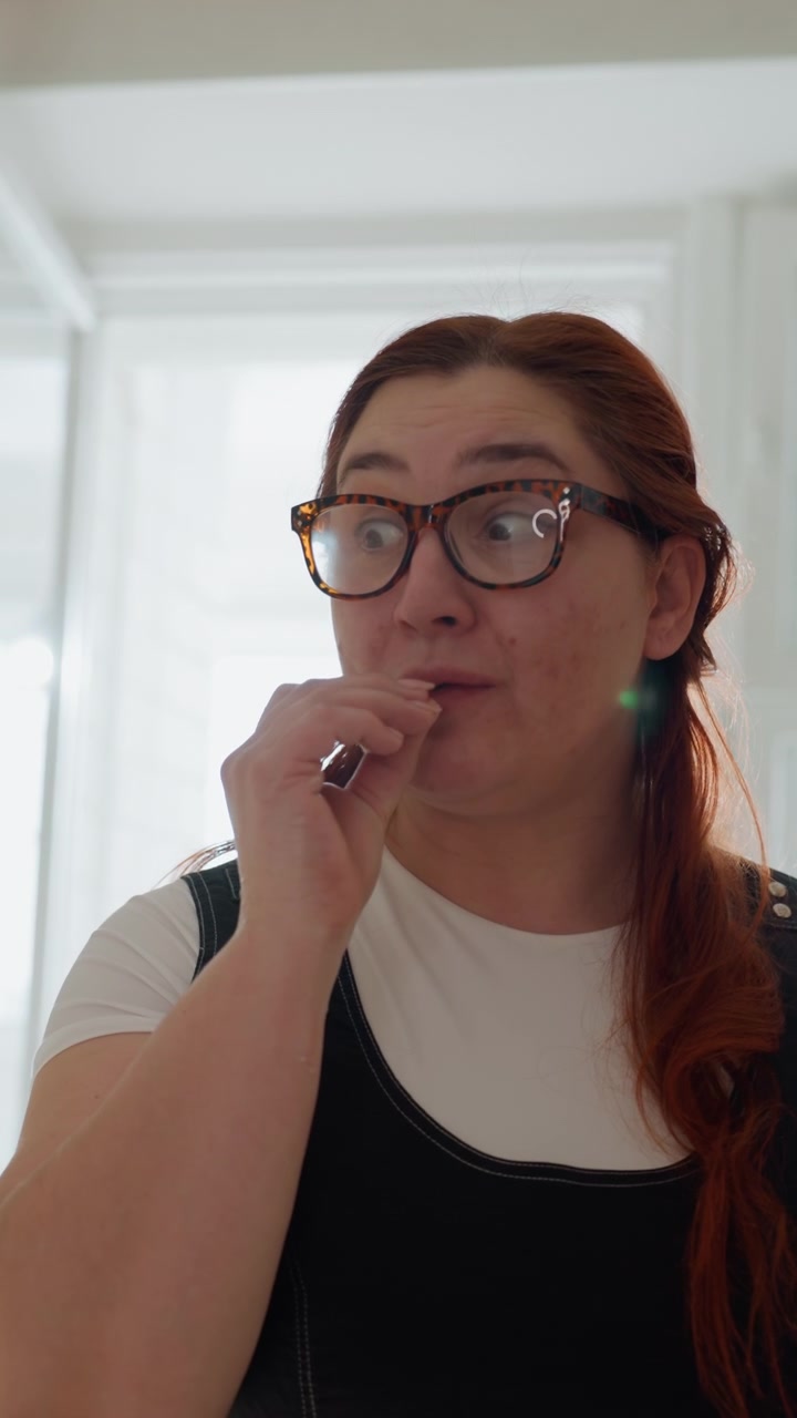 Donna dai capelli rossi con gli occhiali siede in un interno luminoso, facendo piccoli gesti conversazionali e lievi movimenti della testa, con espressione pensierosa, postura calma e morbida luce naturale del giorno.
