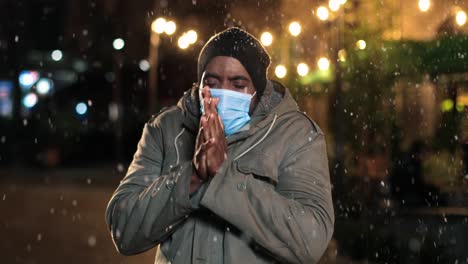 vista de cerca de un hombre afroamericano con máscara facial que tiene frío y trata de calentarse en la calle mientras nieva en navidad