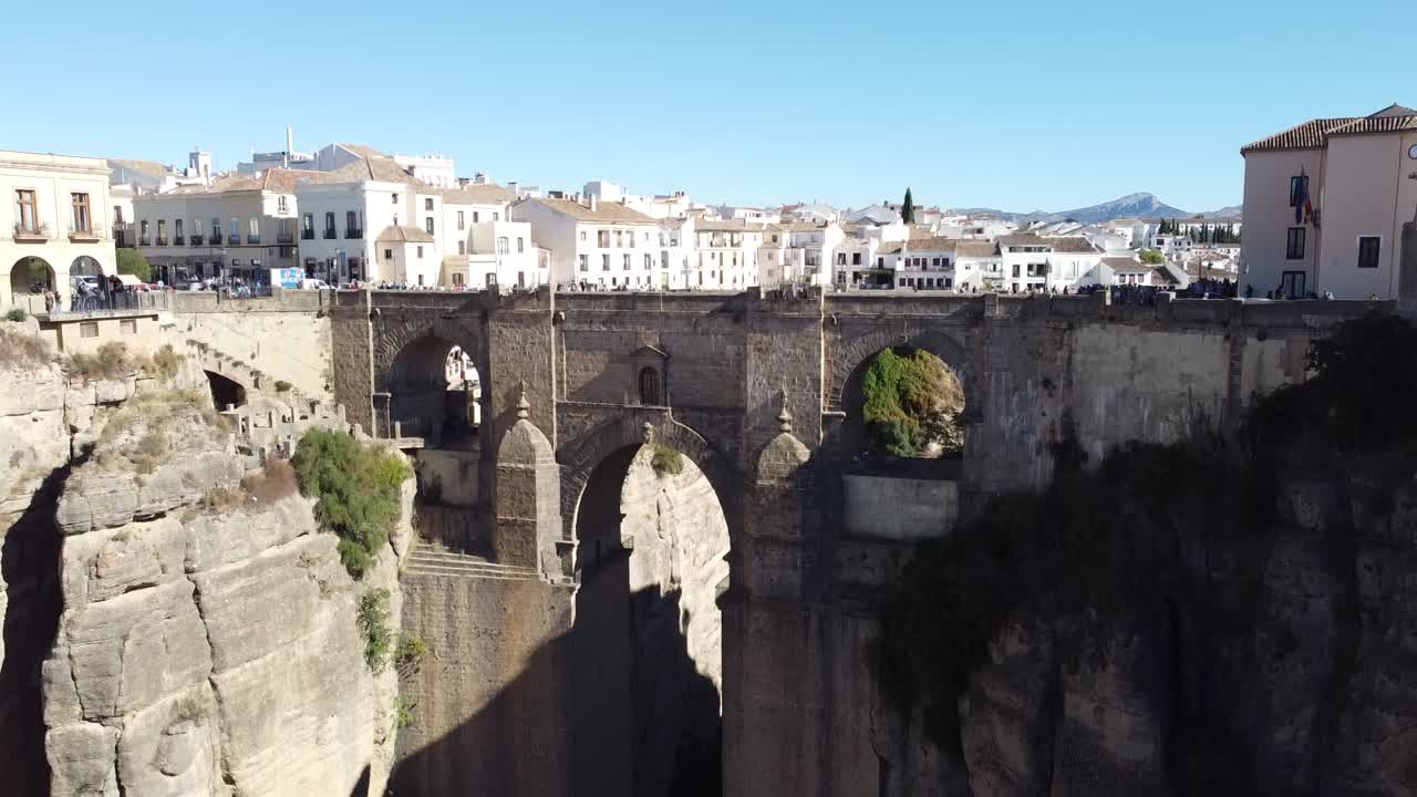 Drone footage showing Puente Nuevo spanning deep gorge with vehicles and people crossing, surrounded by traditional white buildings and dramatic rocky cliffs under clear daylight