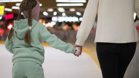 close view of a young girl holding her mom's hand as they skate together on an ice rink, the girl is wearing a mint green outfit with braided hair