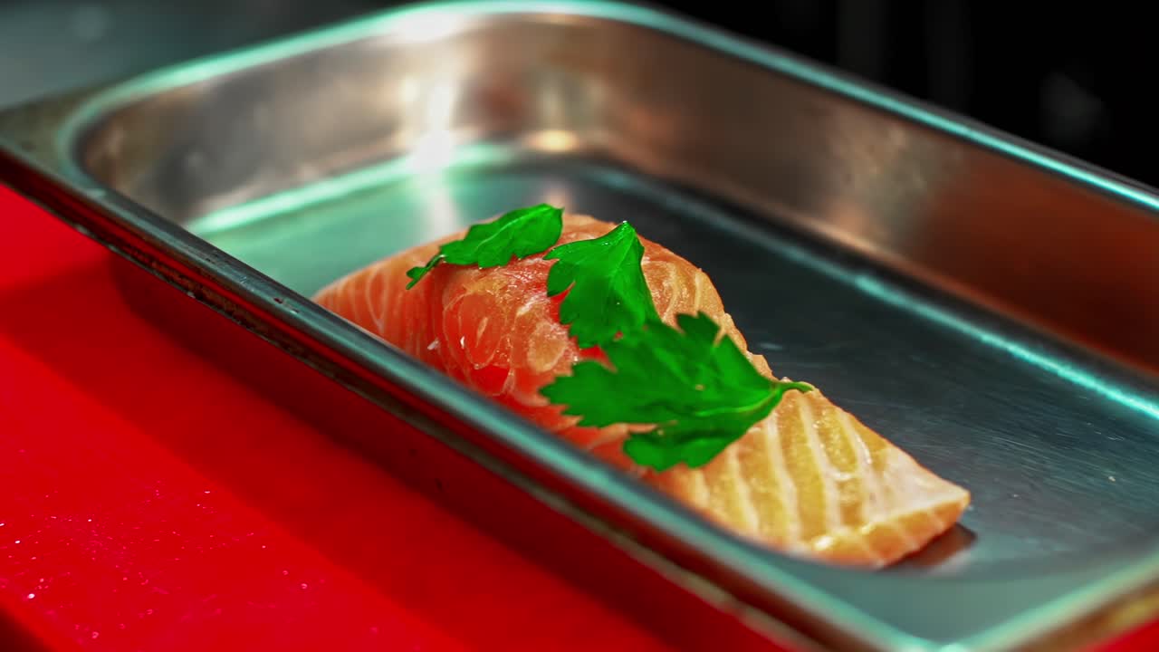 In a professional restaurant kitchen, a chef's hands carefully place a fresh green parsley on a raw salmon fillet in a steel pan, preparing it for cooking a gourmet seafood dish