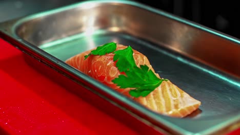 In a professional restaurant kitchen, a chef&#039;s hands carefully place a fresh green parsley on a raw salmon fillet in a steel pan, preparing it for cooking a gourmet seafood dish