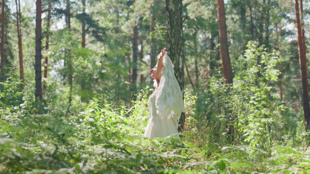 Joyful woman dressed in white turns around smiling warmly under sunlight in lush green forest, raising arms with delight as golden light shines through trees creating feeling of peace