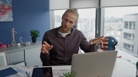 Hombre-Haciendo-Gestos-Llamando-A-La-Computadora-Portátil-En-La-Oficina-De-Cerca.-Video-Chat-De-Diseñador-Serio